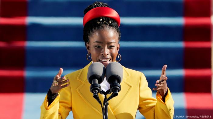 Amanda Gorman, a woman in a bright yellow jacket speaks at a microphone 