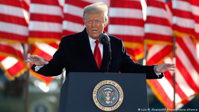 Trump, de terno e gravata vermelha, fala em um púpito. Ao fundo, bandeiras dos EUA. 