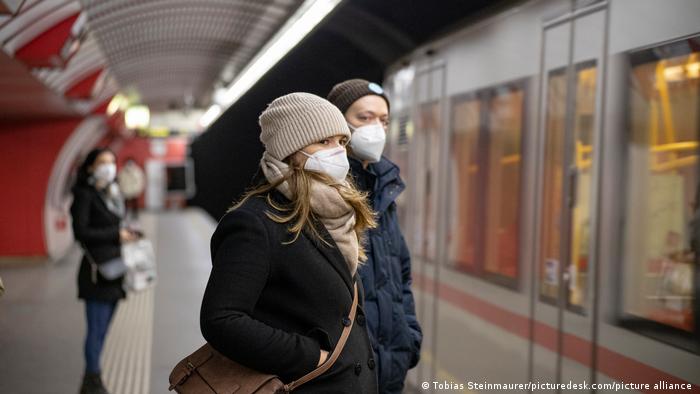 Uma moça e um rapaz de máscara e roupas de inverno aguardam em uma estação de trem. 