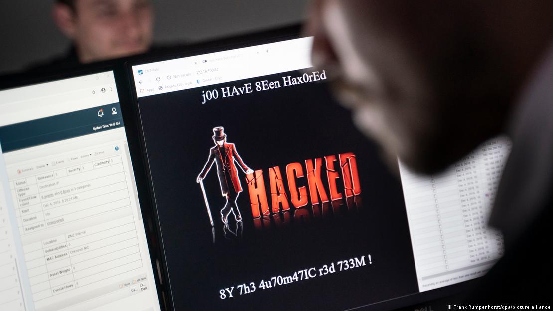 Bilgisayar ekranında "HACKED" yazısı yer alıyor. Ekranın önünde oturan bir kişi, siber saldırı ve zararlı yazılım konulu bir sunuma bakıyor