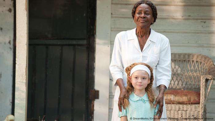 Film still from The Help. A woman has her hands on the should of a young girl. They stand on a porch 