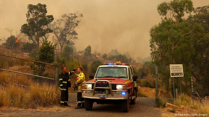 Two firefighters stand next to a fire vehicle as smoke and flames go up behind them. 