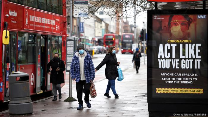 Pessoas caminham com roupas de frio e máscara pela rua. Há um ônibus vermelho à esquerda. À direita, com cartaz da campanha de conscientização sobre o coronavírus.