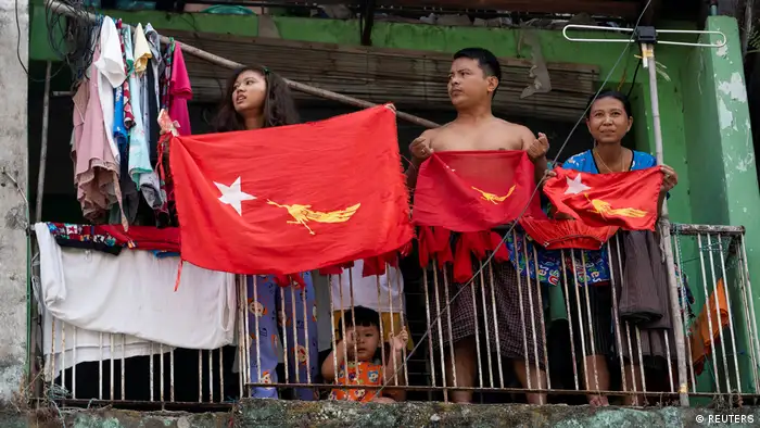Three people holding NLD party flags on a balcony in Yangon, with young child on the ground holding onto the railings