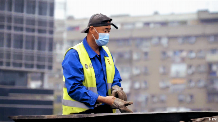 A Chinese worker wearing a mask and yellow high-vis vest pulling on gloves as he works at the train line.
