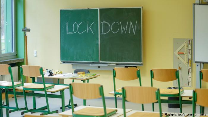 Eine grüne Tafel mit der Aufschrift Lockdown hängt in einem leeren Klassenzimmer an der gelben Wand. An den Schülerpulten davor sind die Stühle hochgestellt