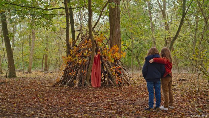 Szene aus Petit Maman: Zwei Kinder stehen im Wald vor einer Hütte aus Stöcken