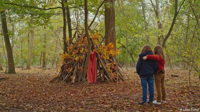 Szene aus Petit Maman: Zwei Kinder stehen im Wald vor einer Hütte aus Stöcken