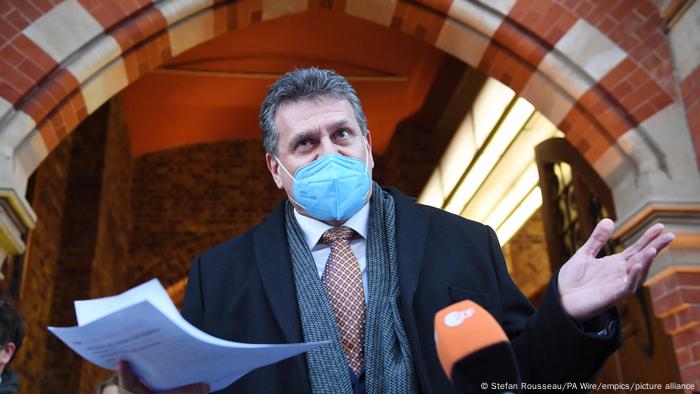 Maros Sefcovic wearing a face mask, standing under a stone arch and holding some papers in one hand 
