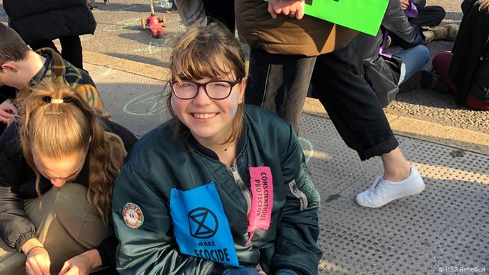 Dos mujeres sentadas en el suelo en una protesta medioambiental. Una de ellas sonríe a la cámara y lleva una chaqueta con un parche de “Extinction Rebellion”.