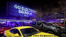 Ein Auto steht auf dem Werksgelände vor einem Gebäude auf das eine Lichtprojektion mit der Aufschrift Go Electric - go Köln geworfen wird. Der Automobilhersteller Ford will seinen weltweit ersten vollelektrischen Kleinwagen in Köln bauen.