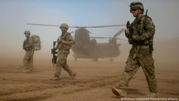 Three armed men walk amid dust with a helicopter in the background
