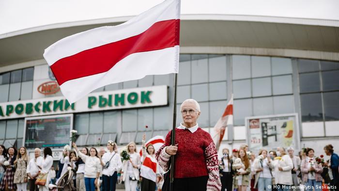 Litauen Ausstellung The Future of Belarus, Fueled by Women: Eine weibliche Demonstrantin gehobenen Alters im roten Pullover hält auf einer Demonstration die weißrussische Flagge in die Luft. Festgehalten wurde der Moment von der Fotografin Viyaleta Sauchyts