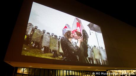 This image shows a photograph projected on the outer walls of the MO museum. The photograph shows two women protesters kissing under the Belarusian flag.