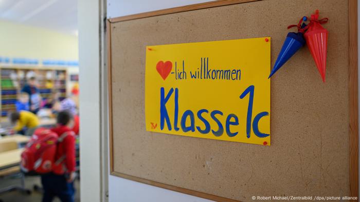 Blick auf die geöffnete Tür und einen Klassenraum dahinter, in dem verschwommen Kinder zu erkennen sind, an der Tür hängt ein Schild, auf dem steht Herzlich willkommen. Klasse 1c
