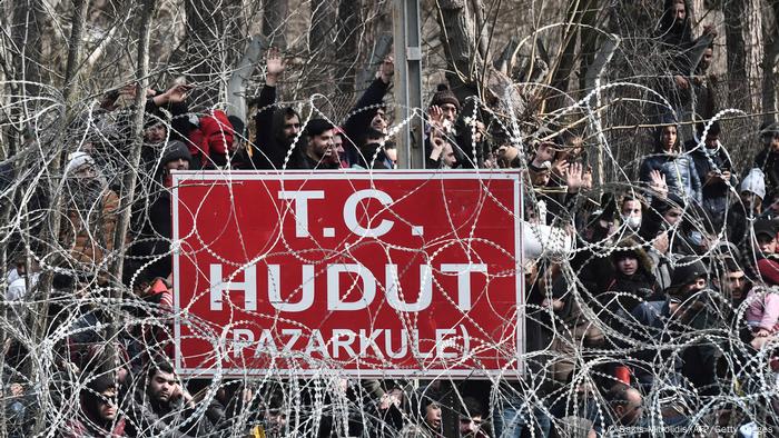 Barbed wire fence with sign in Turkish and migrants waiting on Turkish side