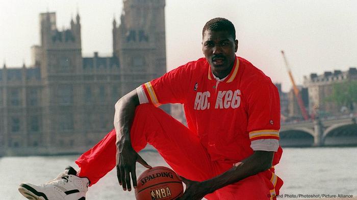 Hakeem Olajuwon sits in a Houston Rockets track suit on a wall by the Thames in front of the Houses of Parliament