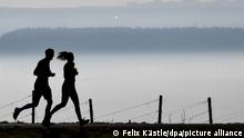 Jogger laufen auf einem Wanderweg in Richtung Meersburg, während im Hintergrund die Stadt Konstanz am Bodensee im Dunst zu sehen ist.