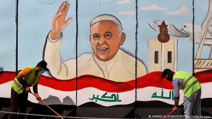  Iraqi municipal workers sweep a street in front of a giant board covered with a painting of Pope Francis.