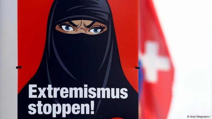 Un drapeau national suisse flotte derrière une affiche contre le port de la burqa.  Il présente le visage d'une femme caricaturale avec un froncement de sourcils voilé et le texte Stop Extremism! 