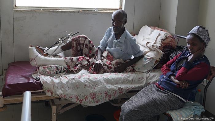 A man with bandaged legs lays on a hospital bed as a woman sits by.