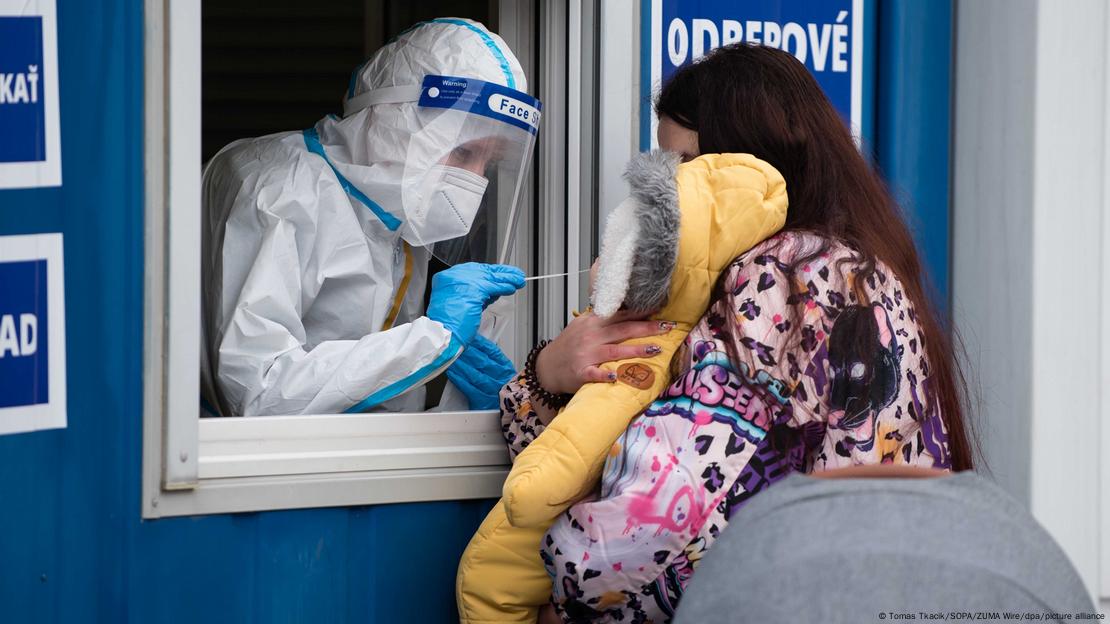 A healthcare worker in protective clothing takes a nose swab sample from a toddler during the Corona mass tests. The child is held by his mother at the window behind which the health worker is standing.