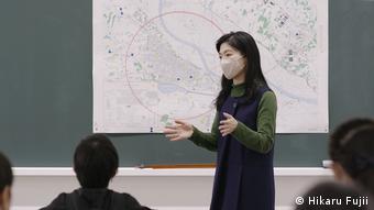 Ausschnitt aus Hikaru Fujii Arbeit The class divided zeigt eine Lehrerin vor einer Landkarte