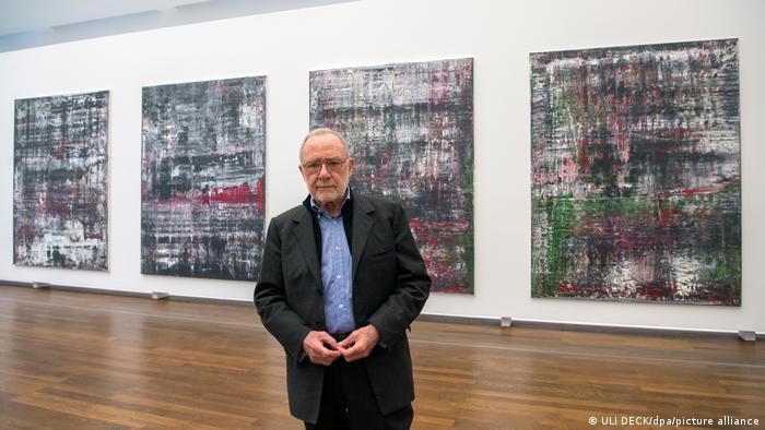 An elderly man stands in front of four abstract paintings