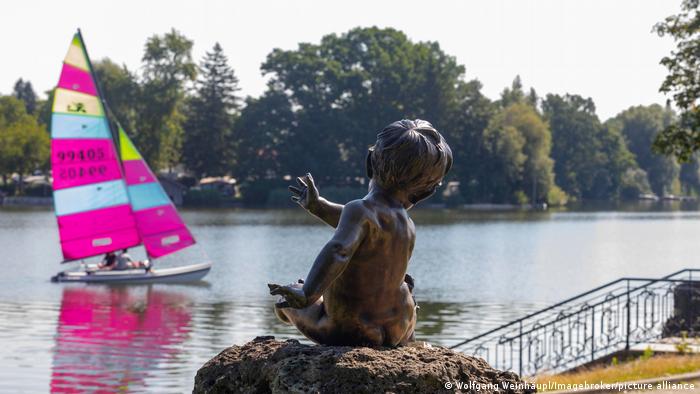 Németország, egy sziklán ülő kisgyermek szobra, aki a tóra néz vitorláshajóval