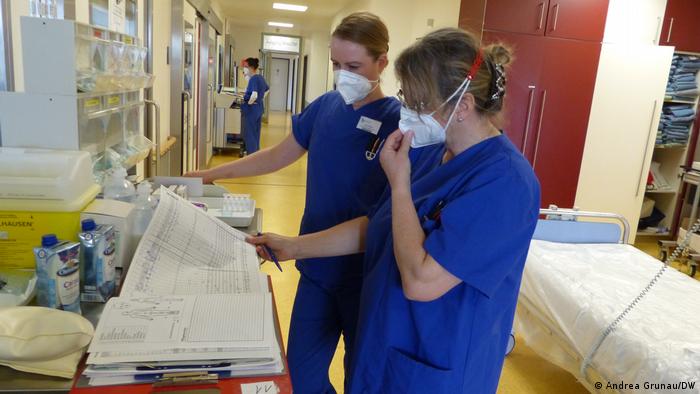 Zwei Frauen in blauer Intensivkleidung mit FFP2-Masken stehen auf einem langen Gang und schauen sich gemeinsam ein Dokument an - die Patientenkurve mit allen Werten