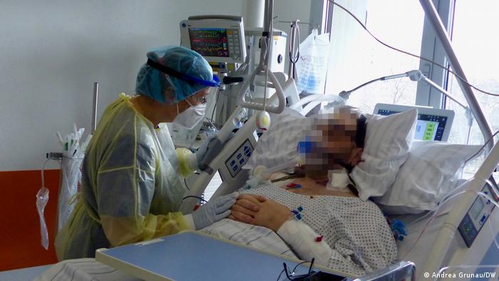 Eine Intensivpflegerin in Schutzkleidung beugt sich über einen Patienten im Krankenbett. Ihre Hand im Handschuh liegt auf seinen Händen, im Hintergrund sind Monitore der Überwachungsgeräte zu sehen
