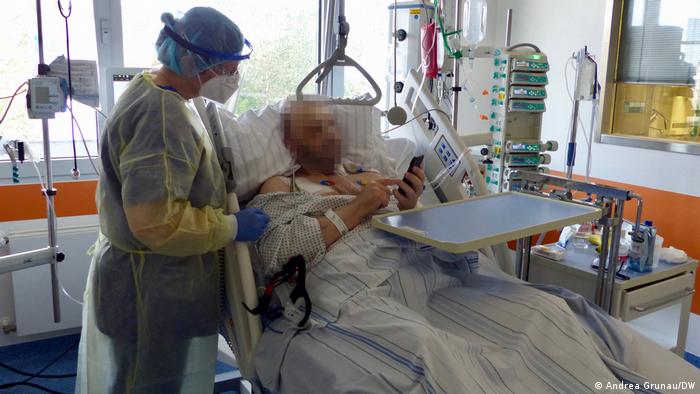 Eine Frau in Krankenhaus-Schutzkleidung steht an einem Krankenbett und schaut zusammen mit dem Patienten auf sein Handy, auf dem er ihr etwas zeigt 