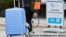 Eine Frau geht mit Rollkoffer am Kuhdamm entlang. Auf dem Schild liest man Corona -Teststation. +++ dpa-Bildfunk +++