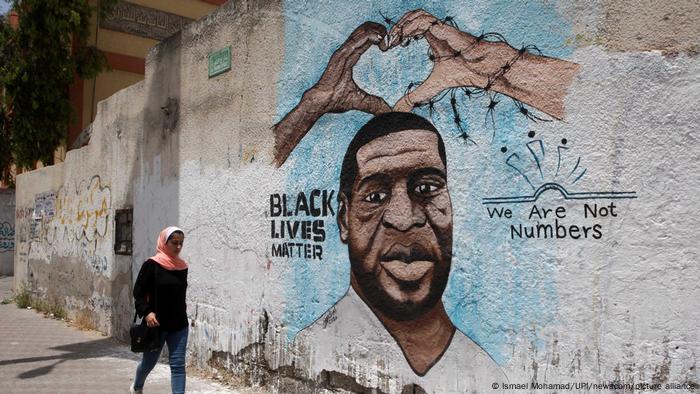 On a sunny day in Gaza City, a woman in a pink headscarf and jeans walks past a portrait of George Floyd, the slogans We Are Not Numbers and Black Lives Matter. 