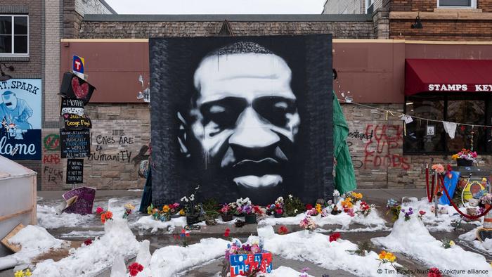 A black and white mural of George Floyd's face. In front of it, mourners have laid bouquets of flowers.