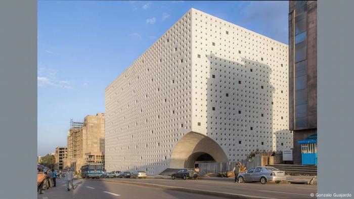 Huge white building with holes in the sides, cars going by on streets in front, and a blue sky.