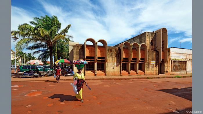 An old bank building wih arched designs on the facade, a palm tree to the left, and women walking across the streets balancing packs on their heads. 