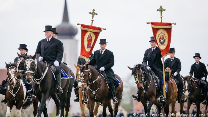Sorb men wearing top hats and black coats, carrying crosses riding on festively adorned horses to mark an Easter tradition.
