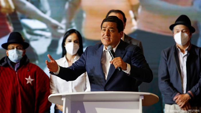 Ecuador Präsidentschaftswahlen - Andres Arauz mit Mikrofon in in der Hand am Rednerpult. Hinter ihm vier Menschen mit Mundschutz.
