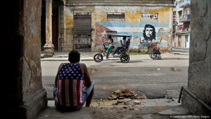 Man wearing a tank top with a design of the US flag sits on the doorstep of a decrepit building In Havana and watching a riksha drive by