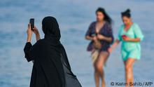 An Emirati woman uses her phone to take a picture as women in bathing suits walk past along a beach in the Gulf Emirate of Dubai on June 25, 2020. (Photo by KARIM SAHIB / AFP)