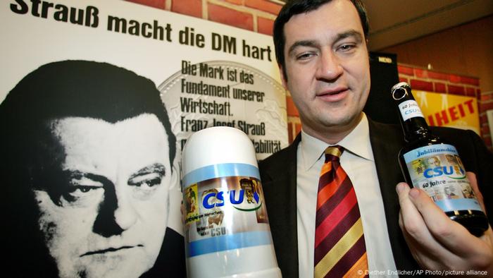 Markus Söder in 2005 holding a beer mug standing in front of a Strauss campaign poster