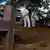 Funcionários trabalham em enterro em cemitério de São Paulo, parentes da vítima observam. Em primeiro plano, uma cruz branca e rosa coberta por ladrilhos. Taxa de mortalidade por grupo de 100 mil habitantes subiu para 181,5 no Brasil.