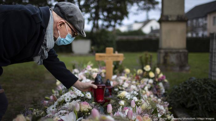An elderly man in a mask places a candle on a fresh grave covered in flowers.