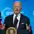 Biden, de terno escuro e gravata vermelha, fala ao microfone. Ao fundo, uma bandeira dos EUA.