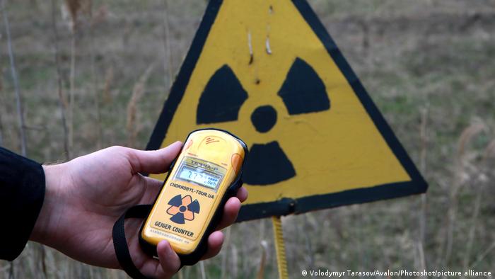 Eine Person hält einen Geigerzähler vor eine Wiese mit einem Warnschild vor radioaktiver Strahlung 