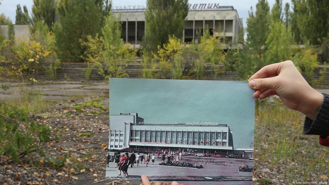 Uma pessoa segura uma foto preto e branco do mesmo prédio que aparece ao fundo e que atualmente está abandonado, na cidade de Pripyat, no norte da Ucrânia, onde ficava a usina nuclear de Chernobyl.