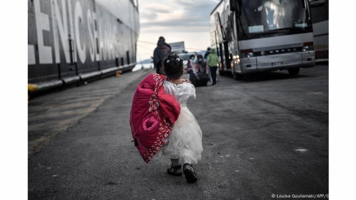 Criança vestindo um vestido branco e um casaco vermelho anda de costas em direção a um ônibus de refugiados na Grécia