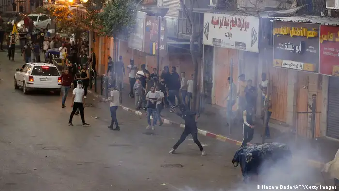 Smoke over a street with many men; one masked man gets ready to hurl an object
