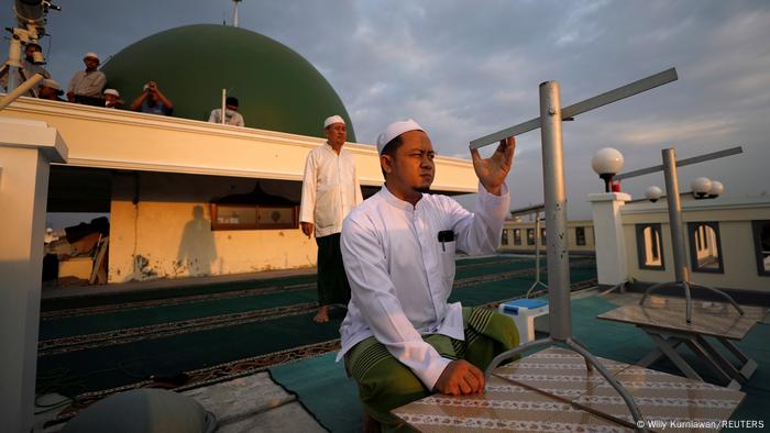 Ein Mann kniet auf dem Dach einer Moschee und blickt entlang eines metallenen Instruments in den Himmel